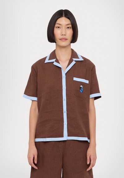 Chemise marron à manches courtes avec un liseré bleu clair, fermeture à boutons et une poche ornée d'un motif de bird brodé en bleu.