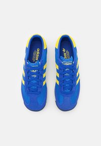 Des chaussures de sport bleu et jaune avec une tige en maille, des accents en daim, trois rayures jaunes et des lacets plats, présentant un design à bout arrondi.