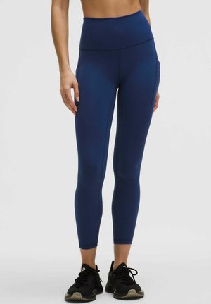 Person iført marineblå højtaljede leggings og sorte sportssko, stående mod en ensartet lys baggrund.