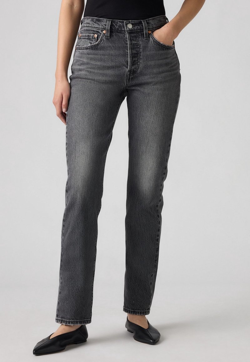 Femme portant un jean noir taille haute, coupe droite, délavé, avec poches avant, associé à des ballerines noires et un haut noir, debout devant un fond gris.