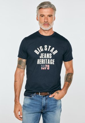 Mężczyzna w średnim wieku z siwymi włosami i brodą, ubrany w czarną koszulkę Big Star Jeans Heritage 1979 oraz niebieskie dżinsy z tatuażami na obu ramionach.