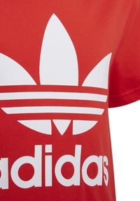 Sarkana kokvilnas T-krekls ar izteiktu baltu trīs svītru logo un trifeles dizainu uz priekšpuses, ar uzdruku „brand” kaut kādā biezuma tekstā zem tā.