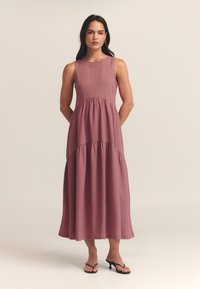 Garais, bezpiedurkņu kleita mauve audumā. Tam ir pieguloša augšdaļa un savākta josta, kas pārvēršas līdzenā svārku daļā. Valkāta ar melniem sandalēm.