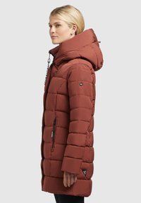 Red puffer coat with a hood, featuring a quilted design, zipper pockets, and a branded badge on the sleeve. Soft texture, mid-length.