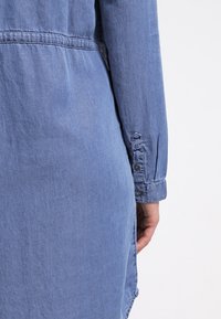 Denimová košile světle modré barvy, s dlouhými rukávy, knoflíkovými manžetami a volným střihem. Obsahuje šev v pase a zakřivený lem.