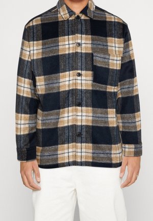 Homme portant une chemise à carreaux marine, beige, gris et blanc avec poche poitrine et un pantalon blanc, sur un fond neutre.