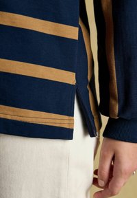 Close-up of a person's hand next to white pants and a navy blue shirt with horizontal beige stripes and a side slit.