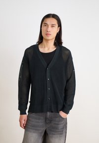 Svart mesh-kardigan med V-hals, långa ärmar och knäppning fram. Har ribbade manschetter och nederkant, bärs över en svart figurnära topp.