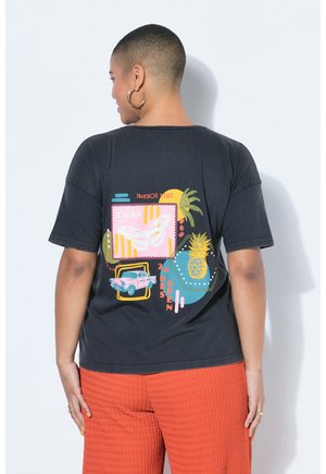 Person med kort hår iført en sort T-shirt med farverigt Cuba-tema print, inklusive en sommerfugl, bil, ananas og tekst, kombineret med orange bukser.