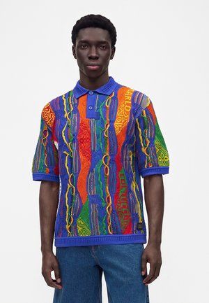 Ung mand stående iført en farverig kortærmet striktrøje med markante lodrette mønstre og blå jeans mod en ensfarvet hvid baggrund.