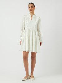 Rochie albă cu accente de volan, decolteu în formă de cheie și mâneci lungi. Decupaje tip eyelet la tiv și la mansete. Asortată cu pantofi de culoare deschisă.