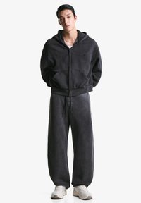 Dunkelgrauer Fleece-Reißverschluss-Hoodie mit großen Taschen, kombiniert mit lockeren, passenden Jogginghosen und hellen Sportschuhen.