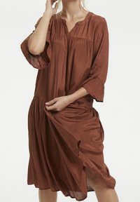 Brun, knælang kjole lavet af glat, flydende stof. Har V-udskæring og 3/4 lange ærmer med samlede detaljer. Casual pasform og drapering.