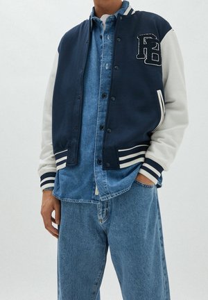 Mężczyzna w granatowo-białej kurtce varsity na niebieskiej koszuli jeansowej i dżinsach, stojący z jedną ręką w kieszeni na jednolitym tle.