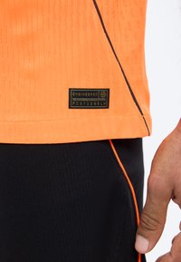 Κοντινό πλάνο ενός ατόμου που φοράει πορτοκαλί πουκάμισο με μαύρη ετικέτα «Engineered» κοντά στο τελείωμα και μαύρο παντελόνι με πορτοκαλί περιγράμματα.