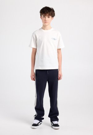 Witte katoenen t-shirt met korte mouwen en een klein bedrukte afbeelding op de linkerborst, gecombineerd met donkerblauwe trainingsbroek met witte strepen aan de zijkant.