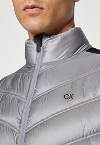 Sivi pufasti kaput s visokim ovratnikom, sadrži zatvaranje na zip, dekorativno šivenje i mali crni "CK" logo na prsima.