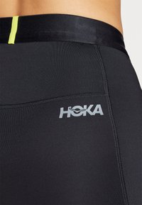 HOKA black athletic leggings with a smooth texture, featuring a slim waistband with a bright yellow accent and logo in silver.