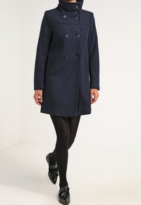 Cappotto blu navy in misto lana con colletto alto, bottoni doppi e due tasche laterali. Vestibilità sartoriale con taglio fino al ginocchio.