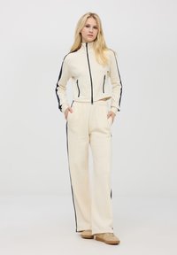 Kremowy zestaw sportowy składający się z krótkiej kurtki z zamkiem błyskawicznym z granatowymi bocznymi paskami oraz pasujących luźnych spodni z elastycznym paskiem.