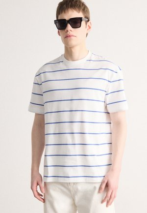 White short-sleeve T-shirt with blue horizontal stripes, round neckline, and relaxed fit. The model wears black rectangular sunglasses.