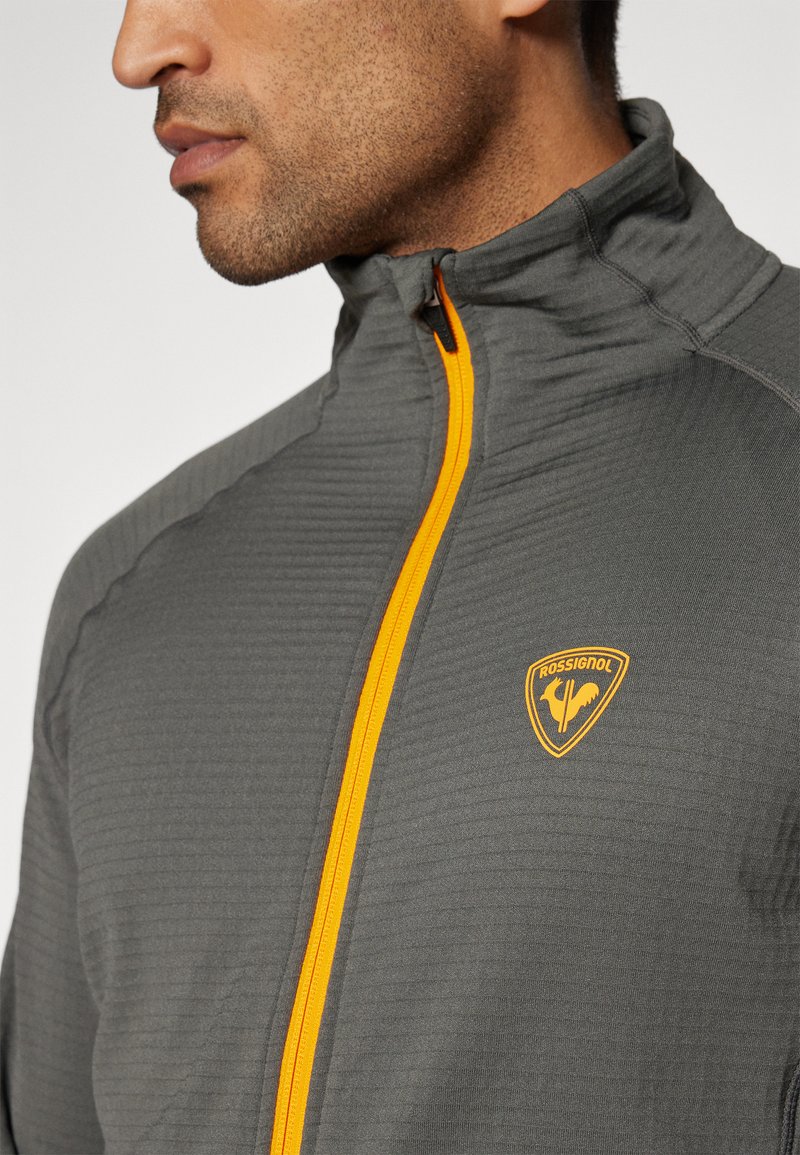 Mężczyzna w szarej bluzie rozpinanej z jasnopomarańczowym zamkiem i logo Rossignol na piersi, zwrócony w lewo, pokazujący żuchwę i szyję.