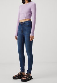 Lila crop-top med långa ärmar, matchat med blå skinny jeans med framsida fickor och knäppning. Svarta slidesandaler.