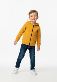 Żółta bluza z kapturem i zamkiem z przodu, noszona na niebieskich dżinsach. Białe trampki, casualowy styl, dziecko stoi z jedną nogą wysuniętą do przodu.