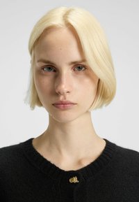 Svart tröja med ribbad halsringning och en guldfärgad knapp. Modellen har kort, rak blont hår och neutral makeup med klar hy.