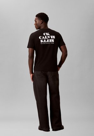 Hombre con camiseta negra Calvin Klein con texto de logo blanco en la parte posterior y vaqueros oscuros, de pie frente a un fondo gris liso.