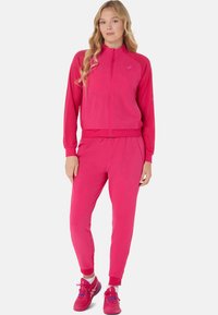Ensemble de sport rose comprenant une veste à fermeture éclair et un pantalon avec cordon de serrage. Comprend des panneaux en maille respirante, des poignets élastiques et des chaussures de sport.