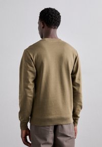 Man med kort lockigt hår, iförd en enfärgad olivgrön långärmad tröja och grå byxor, vänd mot en enfärgad ljusgrå bakgrund.