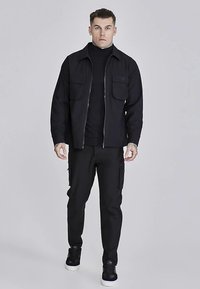 Svart jacka med dragkedja och två framfickor, bärs över en svart polotröja, kombinerad med svarta cargobyxor och svarta sneakers.