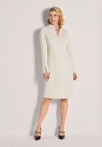 Frau steht vor einer schlichten Wand und trägt ein knie-langes, langärmliges cremfarbenes Kleid mit einem Schlitz-Ausschnitt und schwarze High Heels.