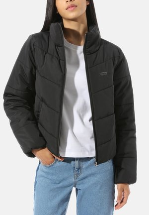 Femme portant une veste matelassée noire sur un t-shirt blanc et un jean bleu, debout avec une main dans la poche et l'autre détendue.