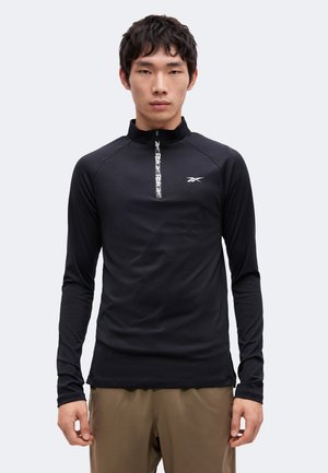 Jeune homme portant un haut de sport noir à manches longues avec une fermeture éclair, un logo blanc et un pantalon kaki, debout devant un fond uni.