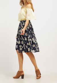 Haut jaune à épaules dénudées avec des manches amples, associé à une jupe florale marine ornée de marguerites blanches, et des sandales à talons ouverts marron.