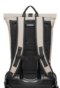 Sac à dos beige avec des sangles noires et un dos en maille. Comprend une poignée supérieure, des sangles réglables et une boucle pour un attachement sécurisé aux bagages.