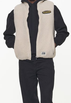 Osoba w beżowej kamizelce polarowej z zamkiem na czarnej bluzie z kapturem i czarnych spodniach, ręce w kieszeniach kamizelki, twarz niewidoczna.