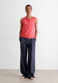 Κοραλί αμάνικο τοπ με ριχτό ντεκολτέ, συνδυασμένο με σκούρους γκρι ψηλόμεσους πλισέ παντελόνες και ανοιχτόχρωμες επίπεδες παπούτσια.