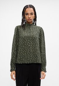 Σκούρο πράσινο φλοράλ μπλουζάκι με μακριά μανίκια και ψηλό γιακά, διακοσμημένο με μικρά λευκά λουλούδια και λεπτομέρειες με κουμπιά στο μπροστά μέρος, φτιαγμένο από ελαφρύ ύφασμα.