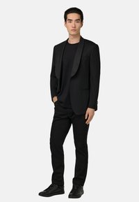 Svart blazer med satinrevers, bärs över en svart t-shirt, i kombination med svarta jeans och svarta sneakers, står framför en enkel bakgrund.