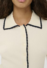 Detajlni pogled na žensko, ki nosi smetanasto ribano pleteno majico z teksturiranim ovratnikom in črnimi valovitimi robovi ob gumbni vrstici.