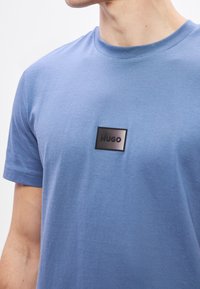 Modré bavlněné tričko s kulatým výstřihem a krátkými rukávy. Na přední straně je obdélníková černá nášivka s nápisem "HUGO".