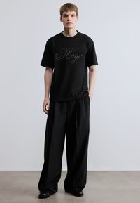 Μαύρο βαμβακερό μπλουζάκι με γραφικό "Hugo" σε στυλ χειρόγραφου, συνδυασμένο με άνετο, φαρδύ μαύρο παντελόνι. Το ύφασμα φαίνεται λείο και μαλακό.