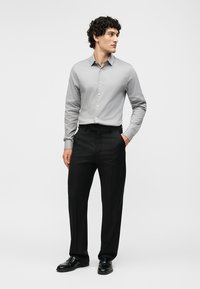 Bărbat cu păr creț purtând o cămașă gri deschis cu nasturi, pantaloni negri eleganți și pantofi negri, stând cu o mână în buzunar.
