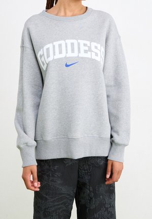Osoba ubrana w jasnoszarą bluzę z napisem „GODDESS” białymi literami oraz małym niebieskim logo Nike, zestawioną z czarnymi, teksturowanymi spodniami.