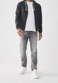 Hombre con sudadera negra con cremallera, camiseta blanca, vaqueros grises y zapatillas blancas de pie contra un fondo blanco.
