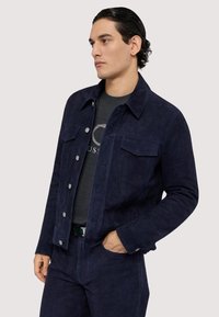 Tmavě modrá semišová bunda se dvěma kapsami na hrudi, stříbrnými knoflíky a klasickým límcem, doplněná tmavě šedým svetrem s kulatým výstřihem pod ní.