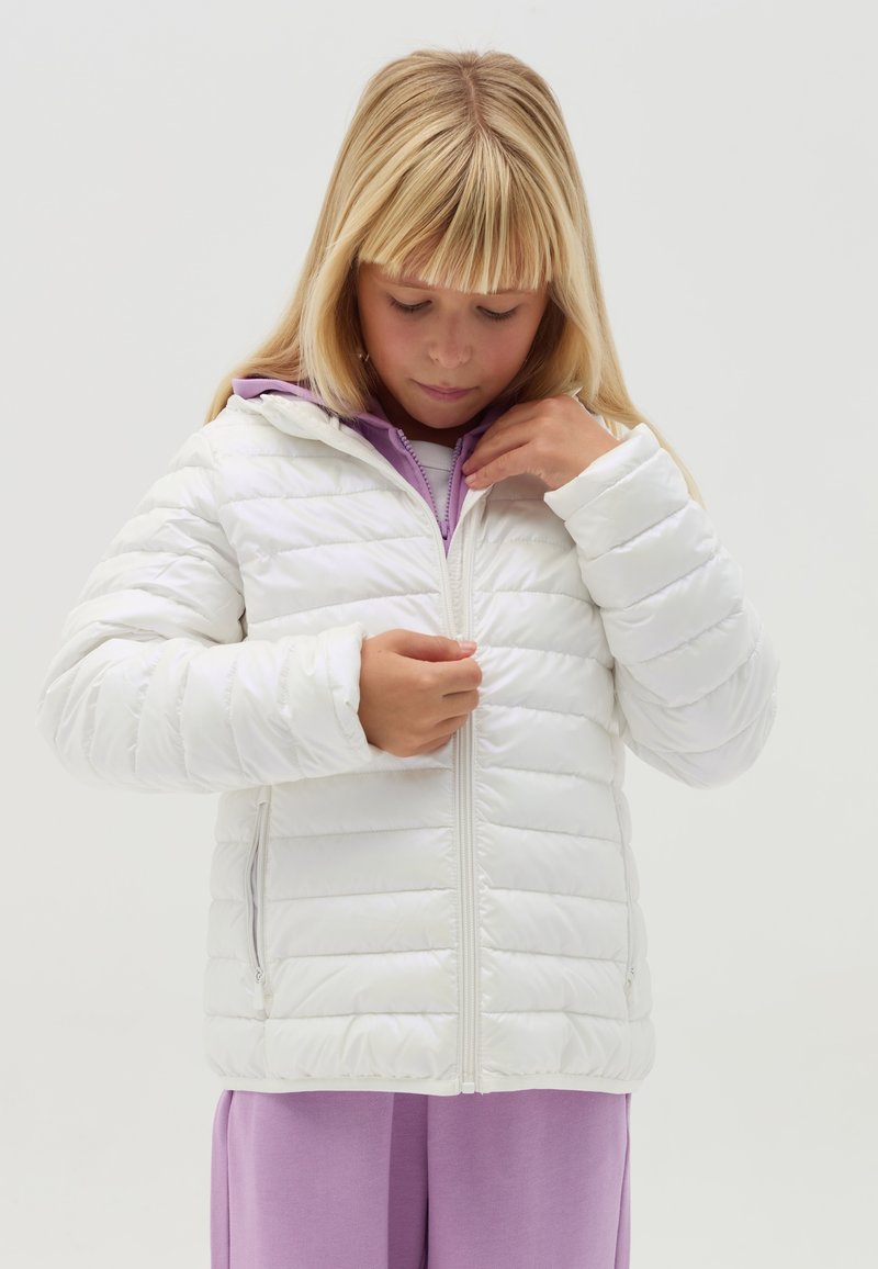 Doudoune blanche avec un design matelassé et une capuche, dotée d'une fermeture éclair à l'avant et de poches latérales, portée sur un haut violet.